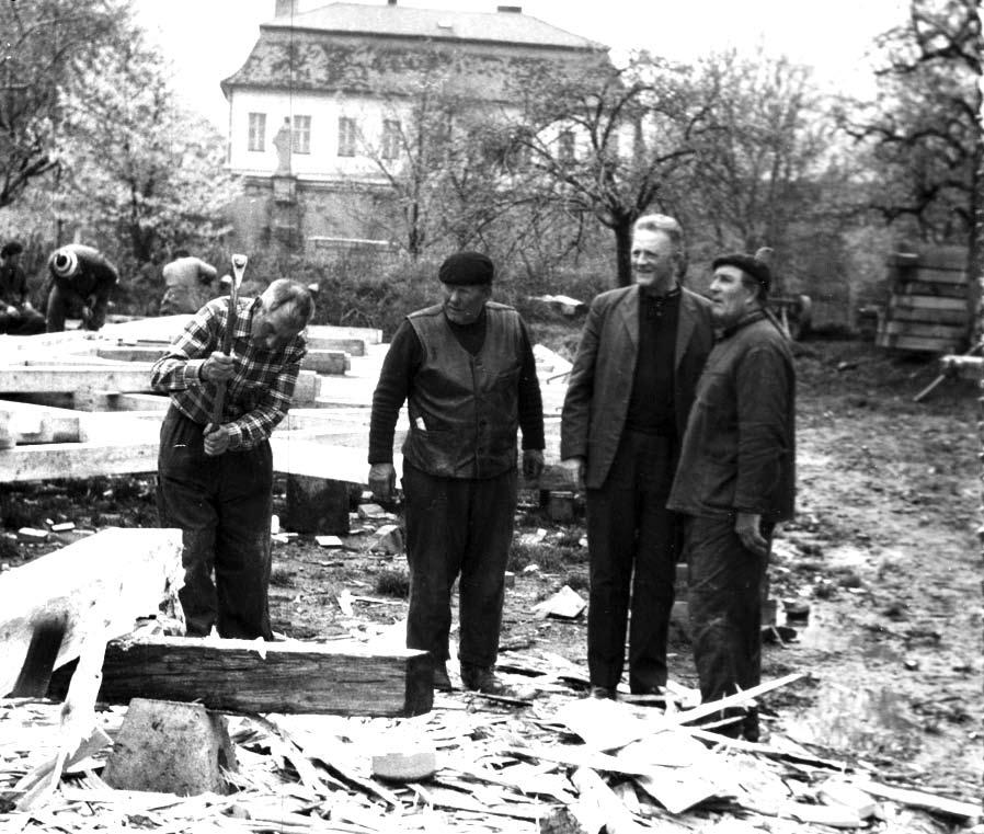 P. Vašíček SJ (uprostřed trojice) přihlíží ručnímu otesávání trámů.