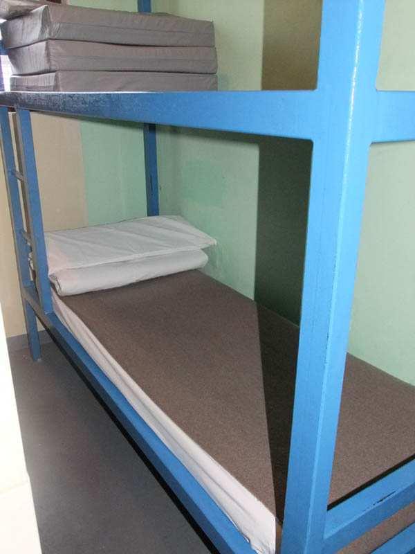 Jedna deka je natažena na prostěradle a je zastlána pod matrací z pohledu ode dveří cely.