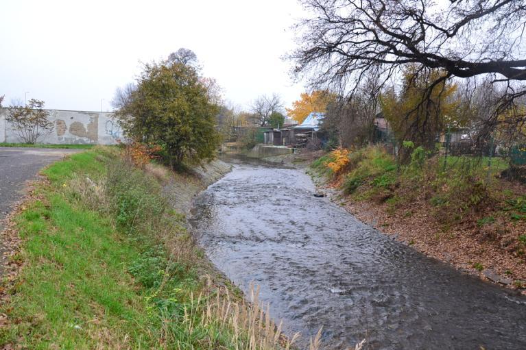 Trasa Koryta zde paralelně vede podél ulice Dukelská. V pravém inundačním území se nachází zahrádkářská kolonie, obytná zástavba stojí v levém inundačním území za paralelně vedoucí silnicí.